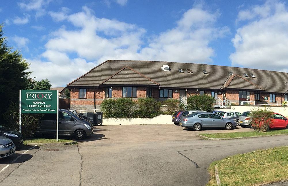 Priory hospital building with sign, cars parked in front, and blue sky.