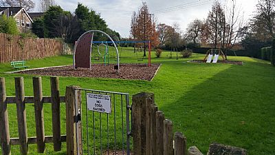 A fenced playground with swings, a slide, and a wooden fence; surrounded by green grass and trees.