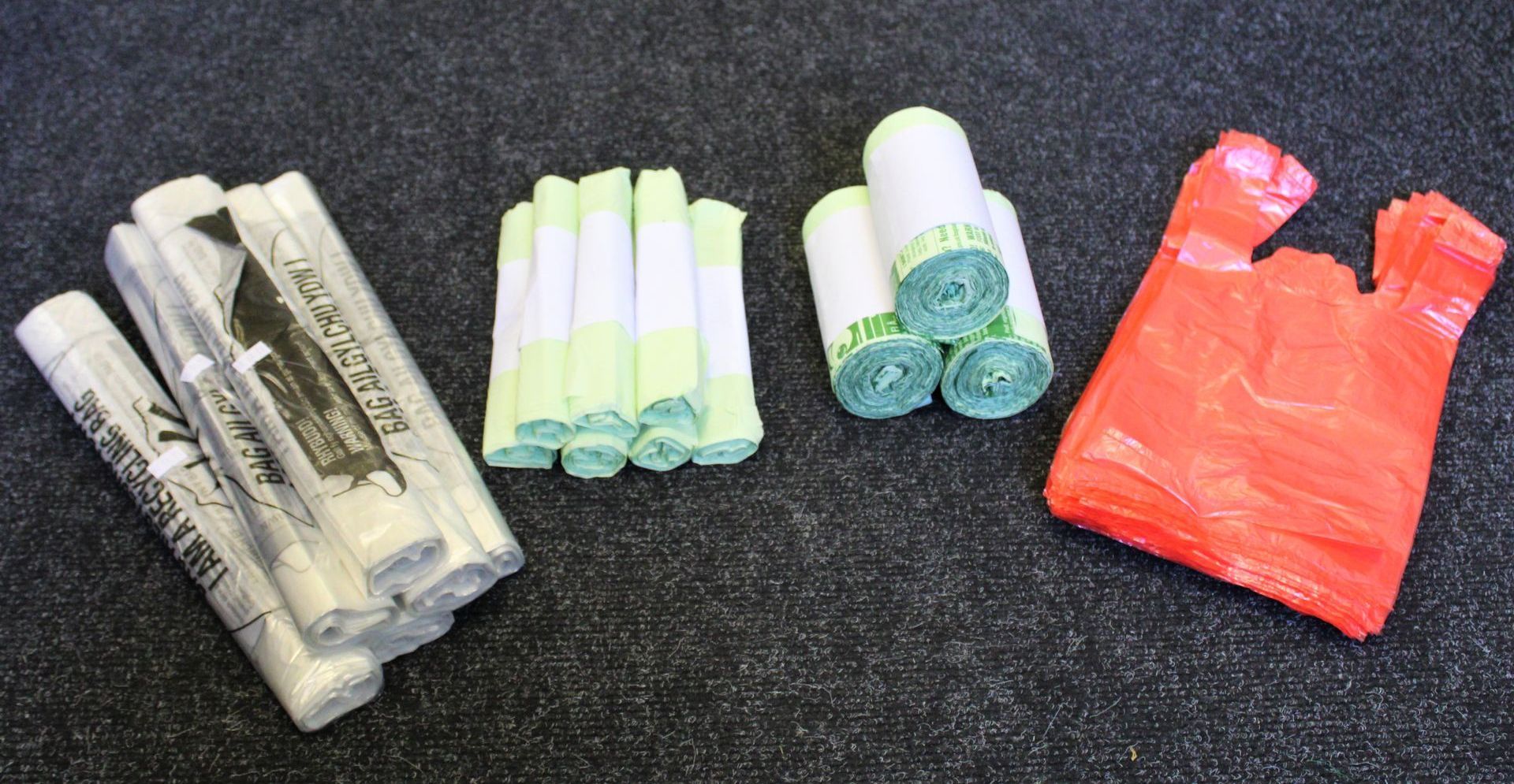 Piles of plastic bag rolls in green and white, a bundle of green bags, and a stack of red plastic bags on a dark surface.