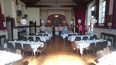 Hall set up for an event, tables with white tablecloths, people mingling, balloons with