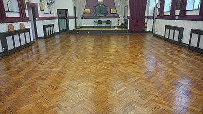 Wooden parquet floor in an empty hall with a stage, curtains, and radiators along the walls.