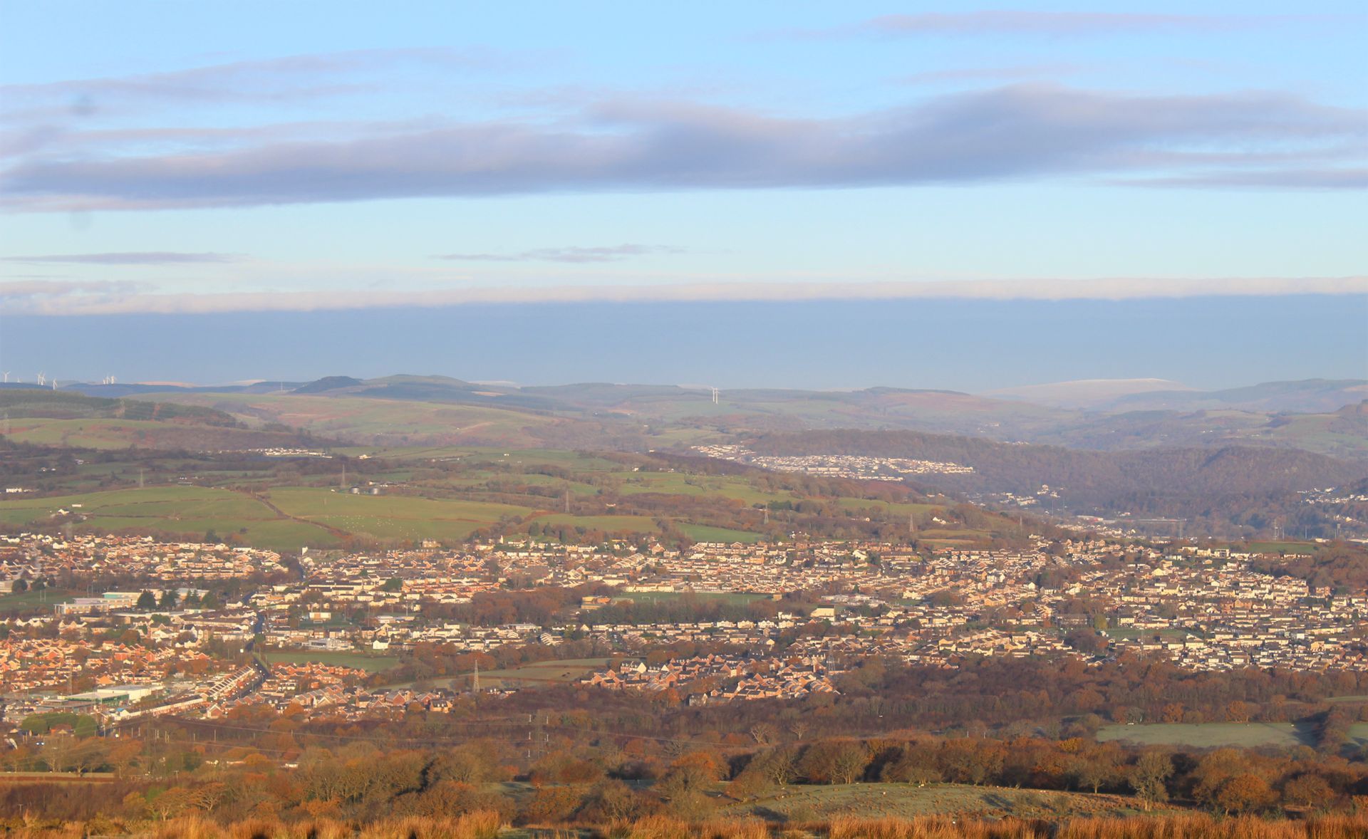 Wide view of a town nestled in a valley, with green hills and a blue sky overhead.