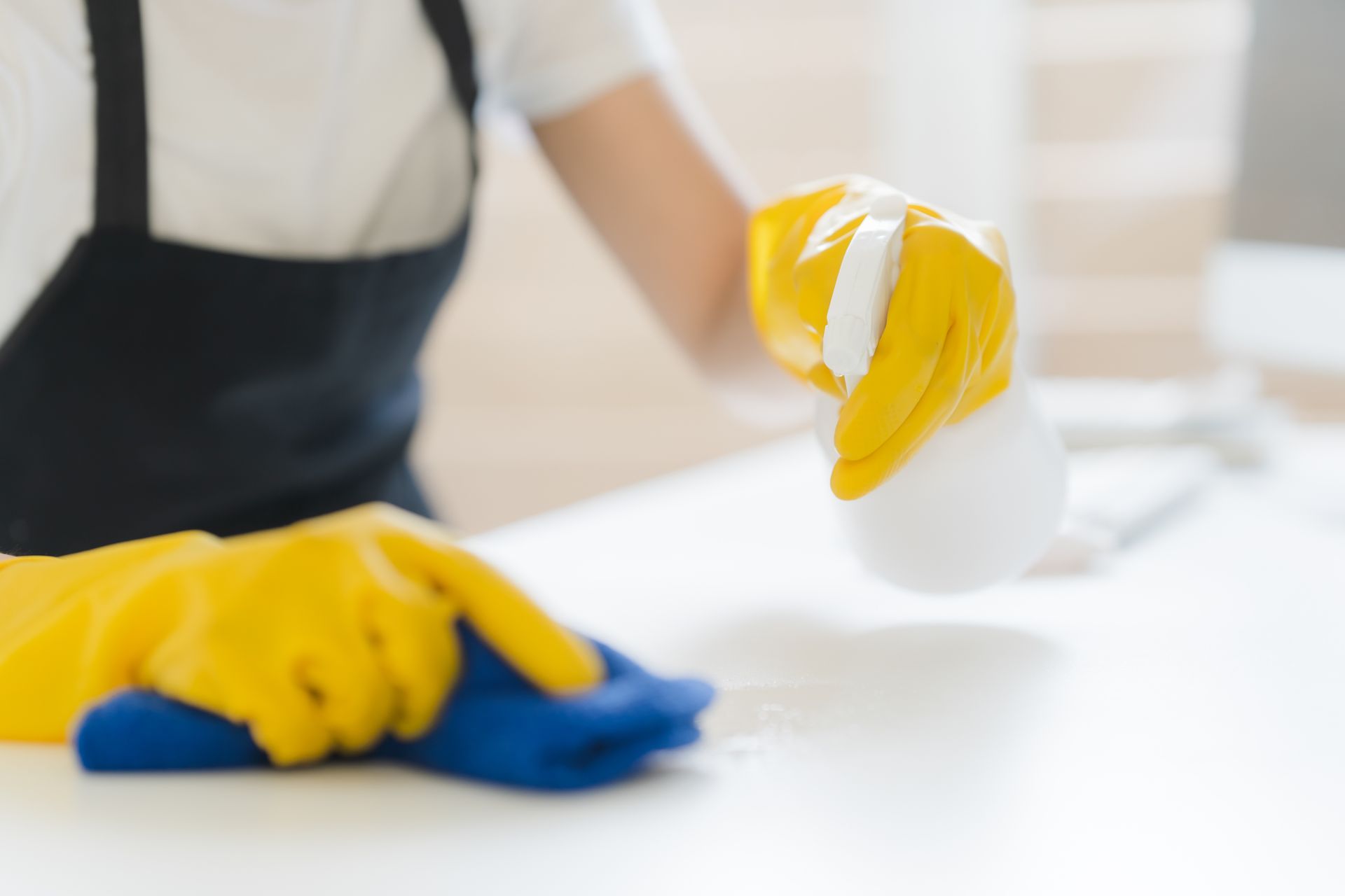 A person wearing yellow gloves is cleaning a counter with a cloth and spray bottle.