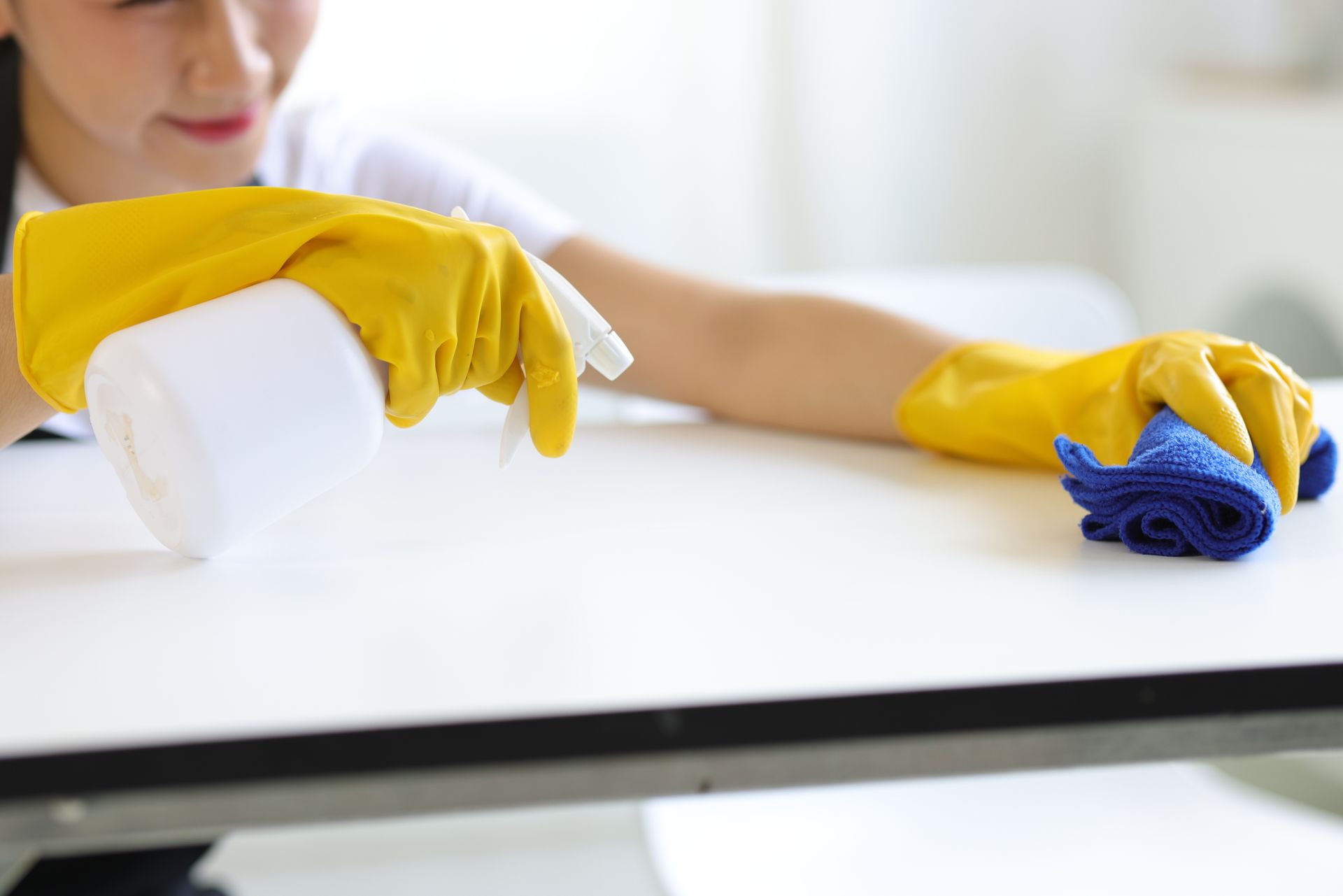 A woman wearing yellow gloves is cleaning a table with a spray bottle and a cloth.
