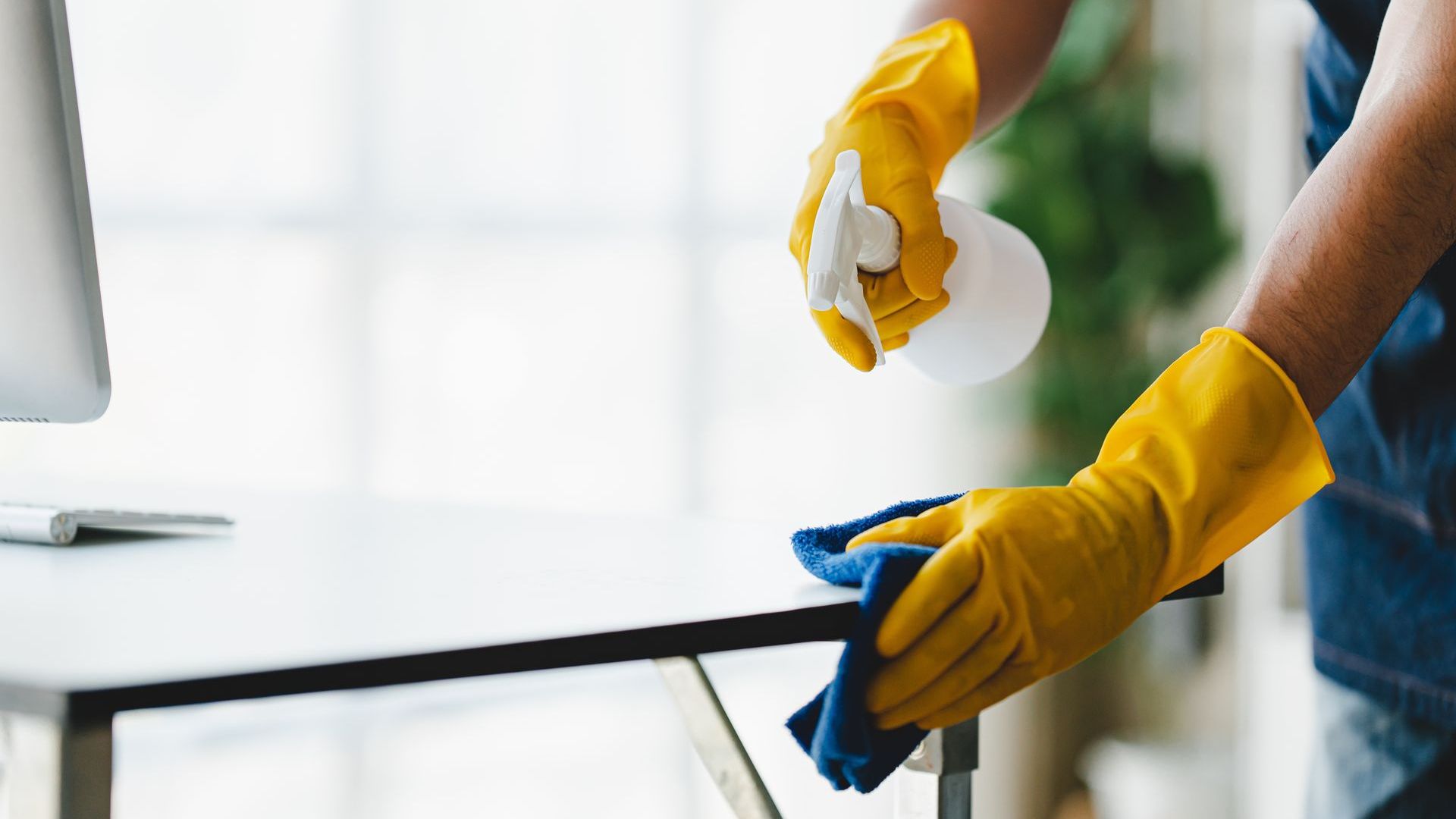 A person wearing yellow gloves is cleaning a desk with a cloth and spray bottle.