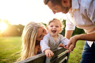 Family smiling outdoors by a wooden fence at sunset, toddler laughing between two adults