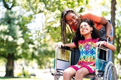 Person in wheelchair smiling with a child in a colorful shirt outdoors in a park
