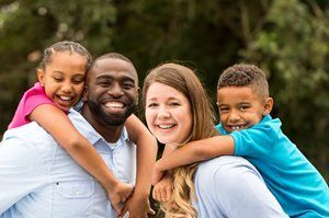 Four smiling people hugging outdoors in a park, with two children leaning on adults' shoulders
