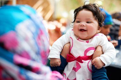Smiling baby in a white outfit with a pink ribbon, sitting in a colorful crowd scene