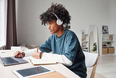 Person wearing headphones studies at a desk with a laptop and notebook in a bright room.