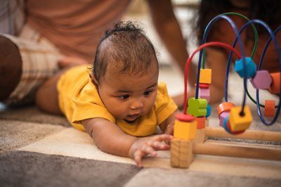 Baby reaching toward a colorful bead maze toy on a rug.