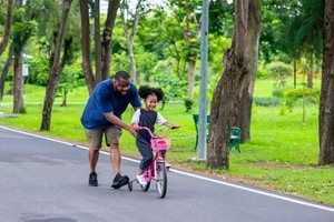 Adult helping child ride a pink bicycle on a tree-lined park path