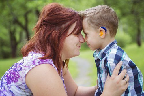Woman and young child touching foreheads outdoors, with greenery in the background