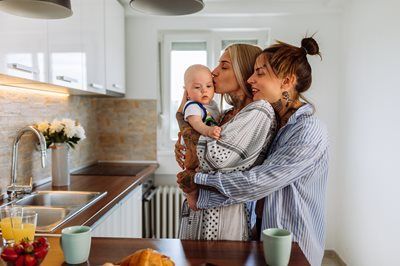 Three people holding a baby in a bright kitchen, standing by a counter with drinks and flowers.