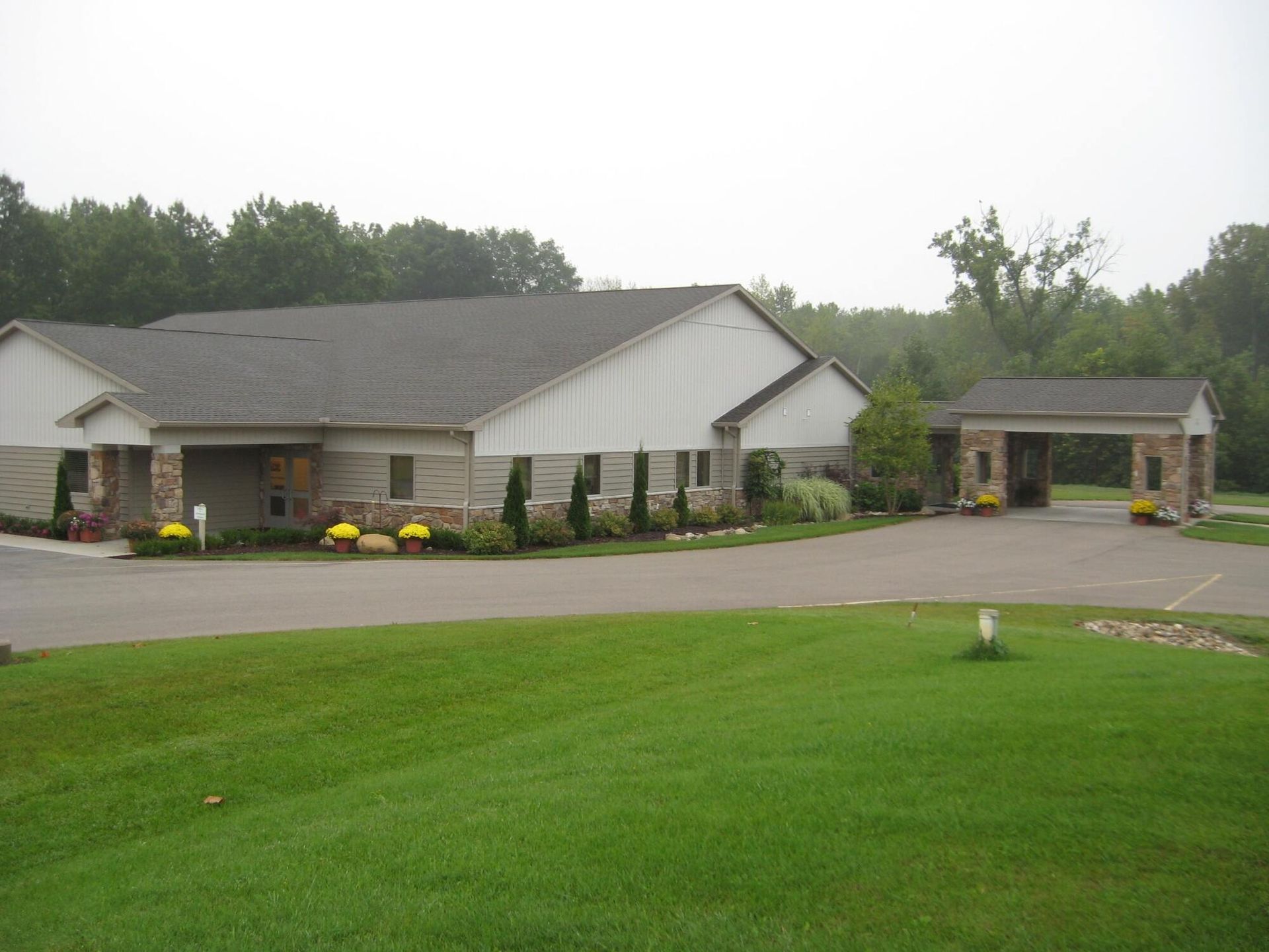 Single-story building with a white facade, flower beds, and a covered driveway on a grassy lawn.