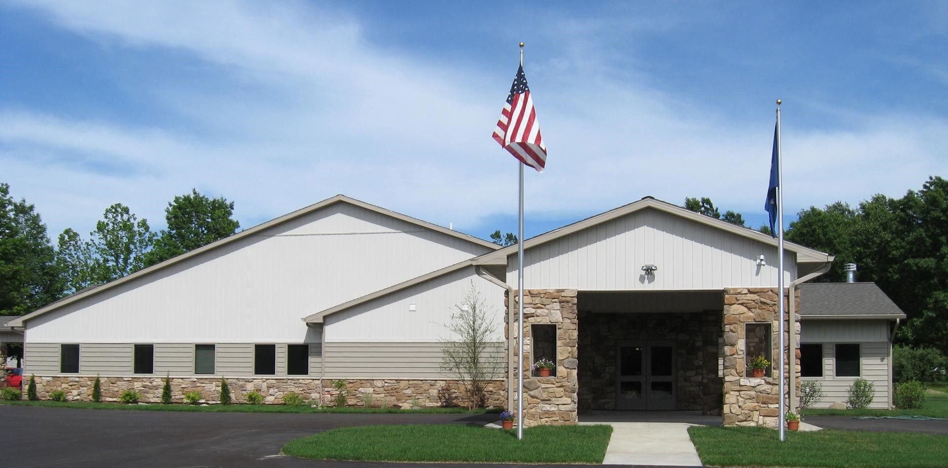 Single-story civic building with stone facade, American flag, and blue flag under a cloudy sky