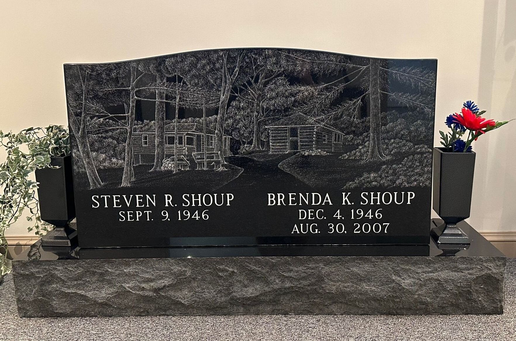 Black granite memorial gravestone with engraved names and dates on a stone base, with flowers at the right side