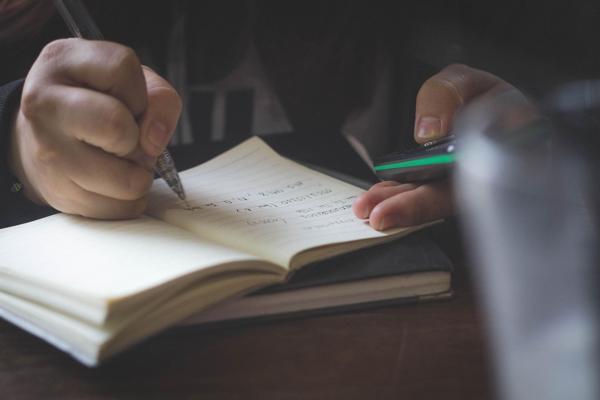 Hands writing in a notebook with a pencil at a desk, with a green pen nearby.