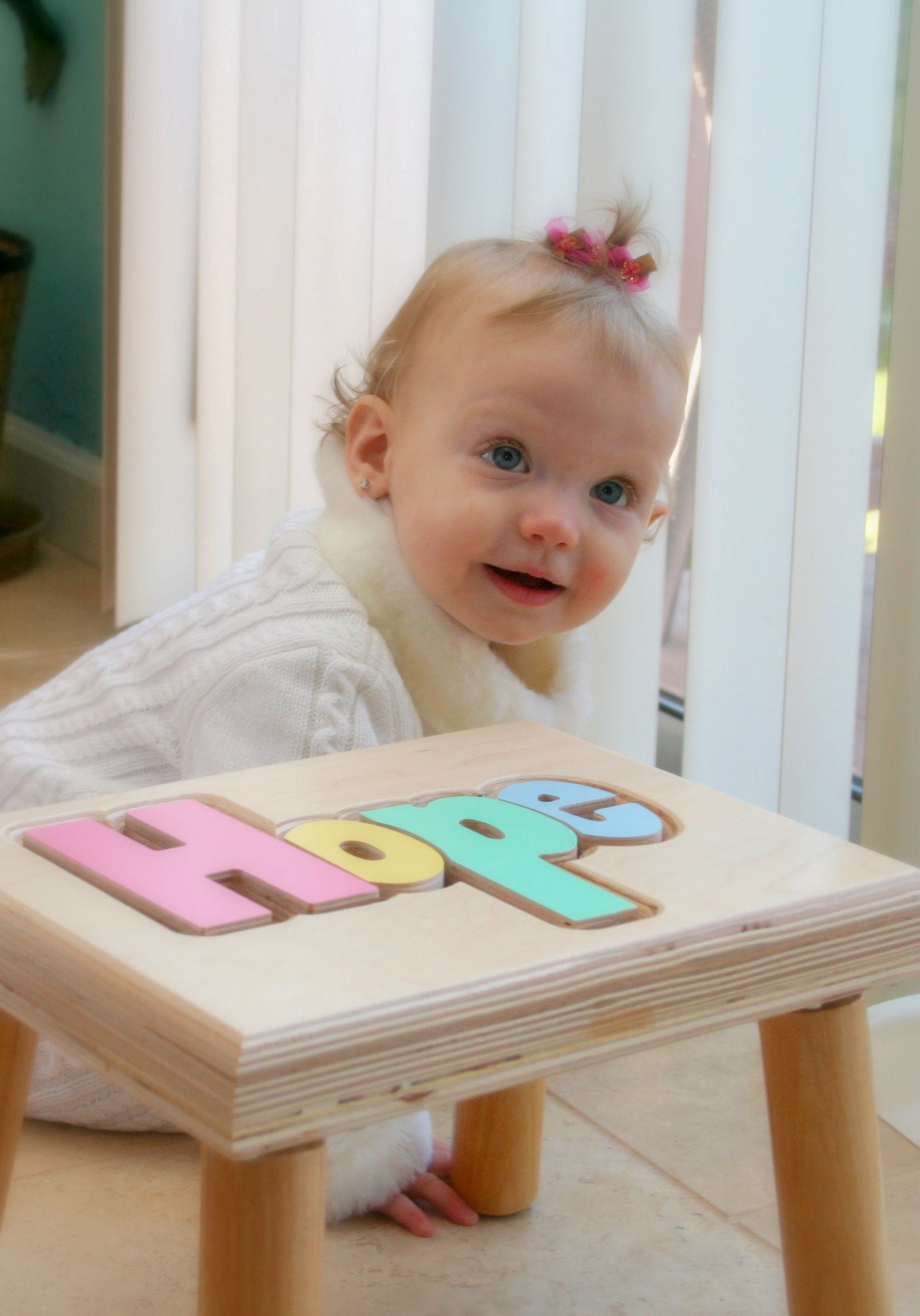 Toddler smiling beside a wooden table with colorful letter blocks in a bright room