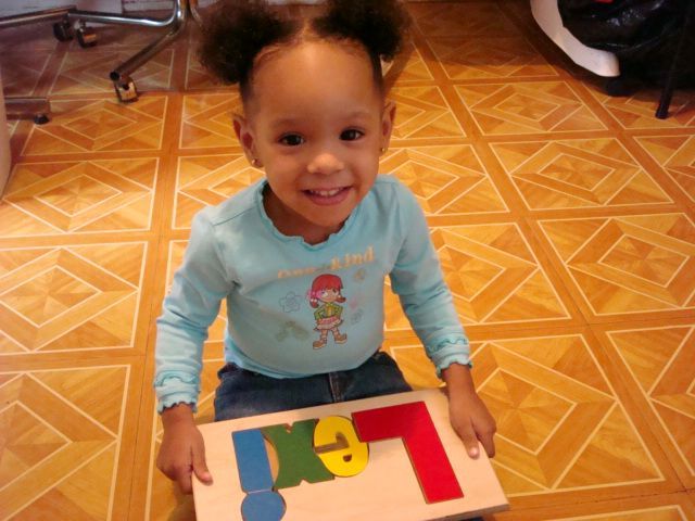 Child smiling on a tiled floor, holding a wooden puzzle board with colorful letter shapes.