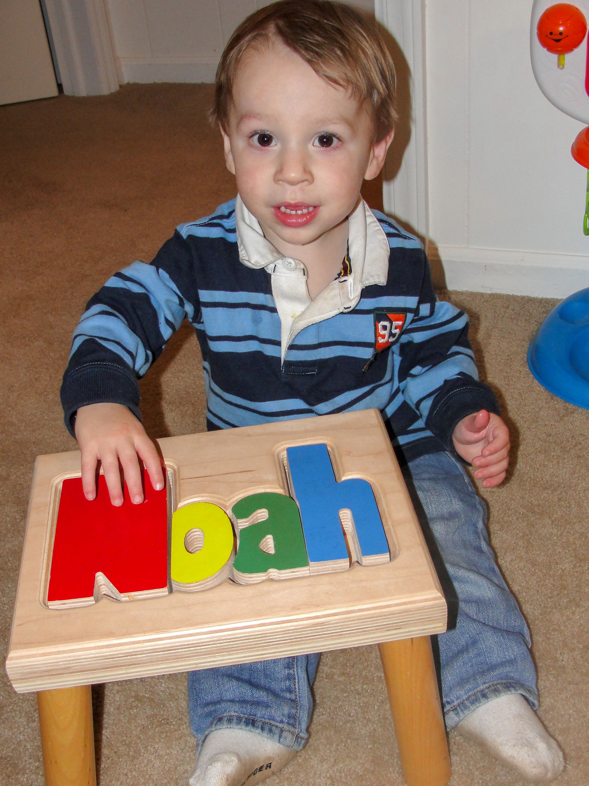 Toddler sitting with colorful wooden name blocks on a small chair, smiling indoors.