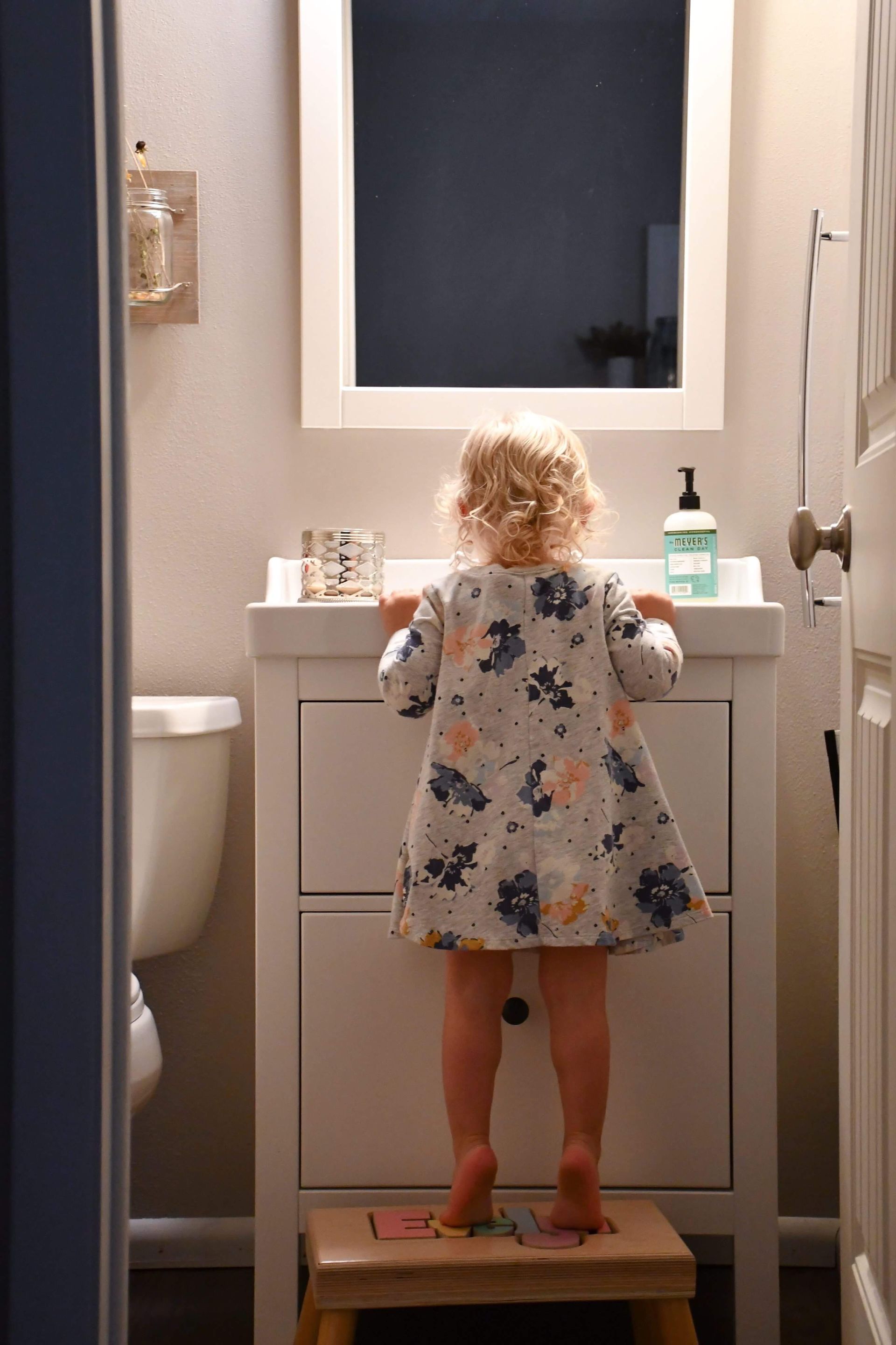 Child standing on a stool at a bathroom sink, looking into the mirror.