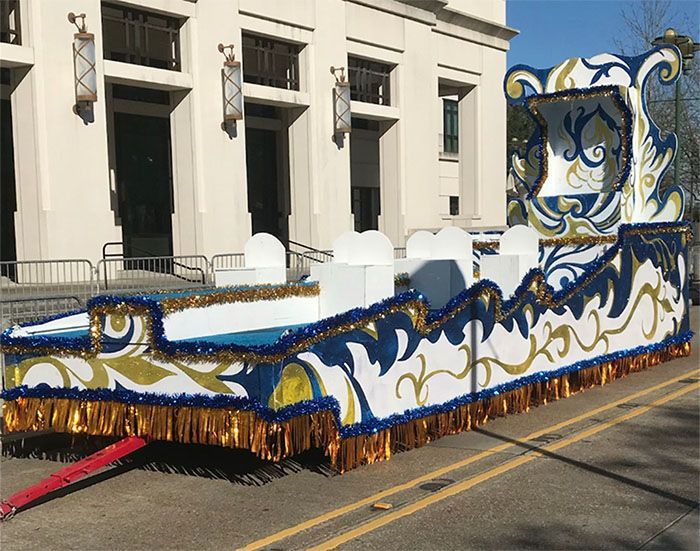 Mardi Gras parade float, white, blue, and gold with ornate scrollwork, parked on street in front of building.