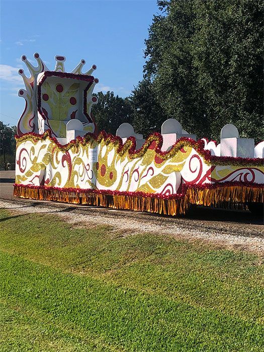 Mardi Gras float on grass; white, gold, and red decorations against a blue sky and trees.