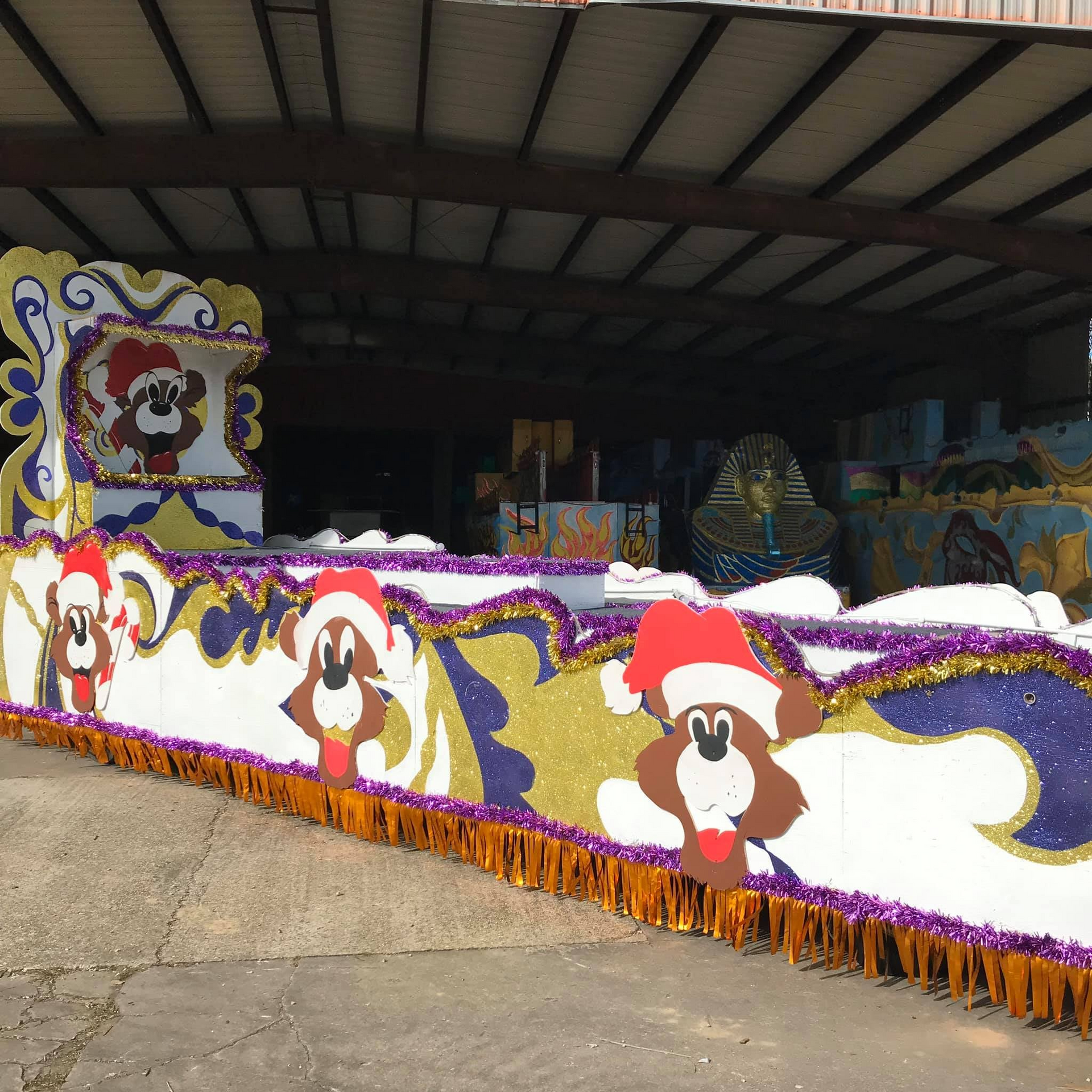 Mardi Gras float with cartoon dog wearing a Santa hat. White, gold, purple, and orange decor under a shed roof.