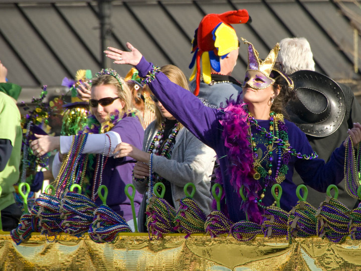 People on a Mardi Gras float throw beads. One woman wears a mask and raises her arm, purple and gold.