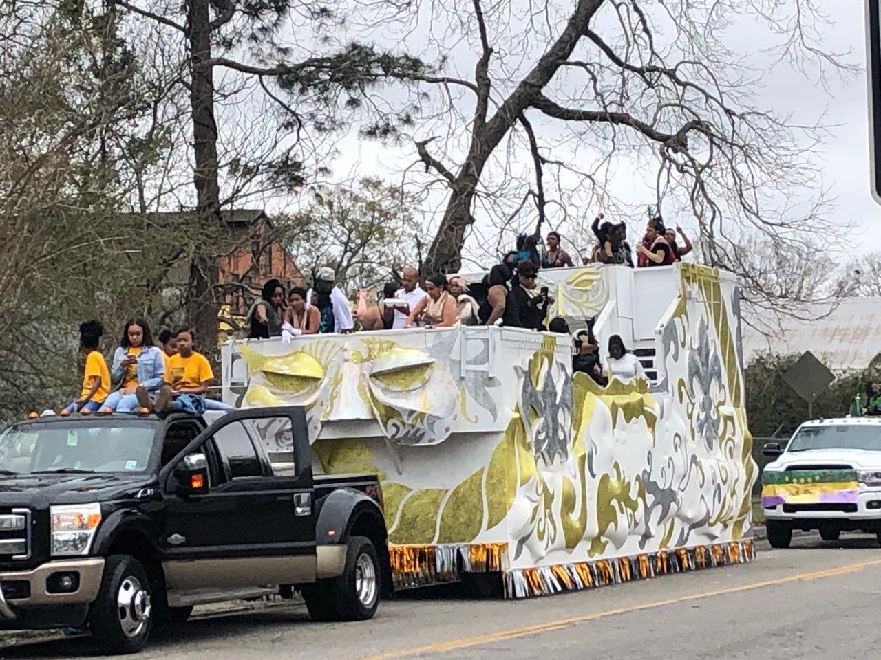 Mardi Gras parade float with people waving. White, gold, and grey design. A black truck is in front.