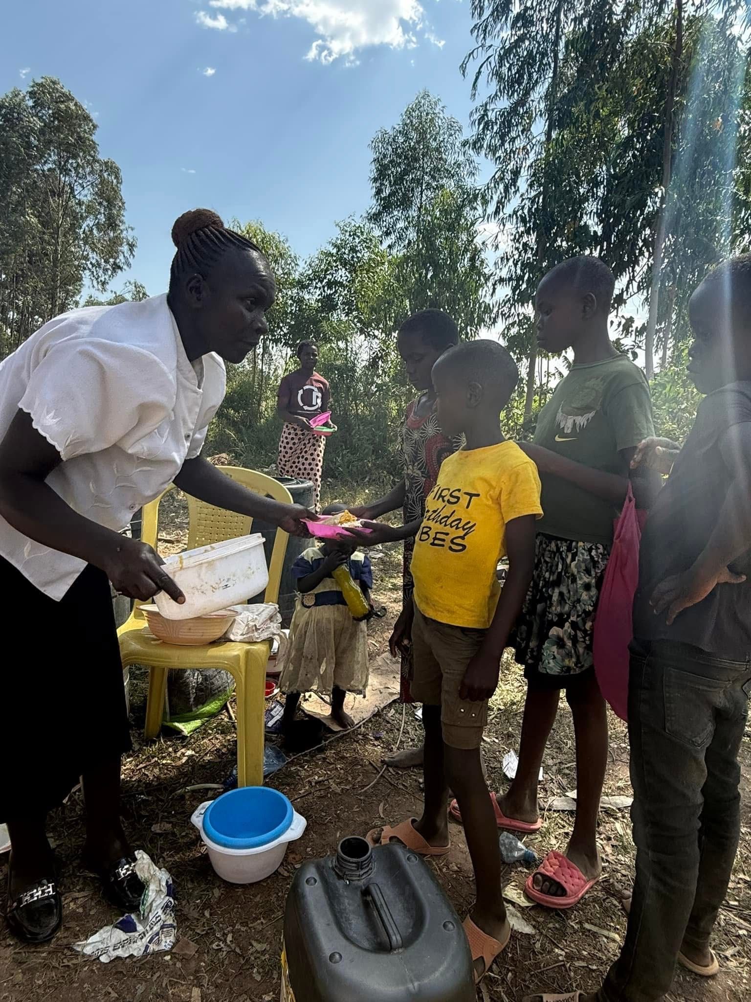 Woman serving food to children outdoors. Yellow shirt. Sunny, trees.