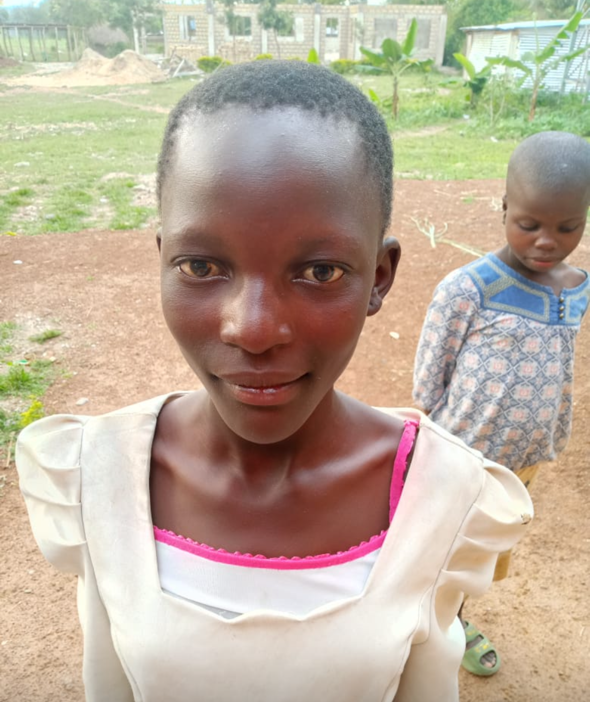 A young Black girl smiles, wearing a white dress with pink trim. A child stands behind her. Outdoors.