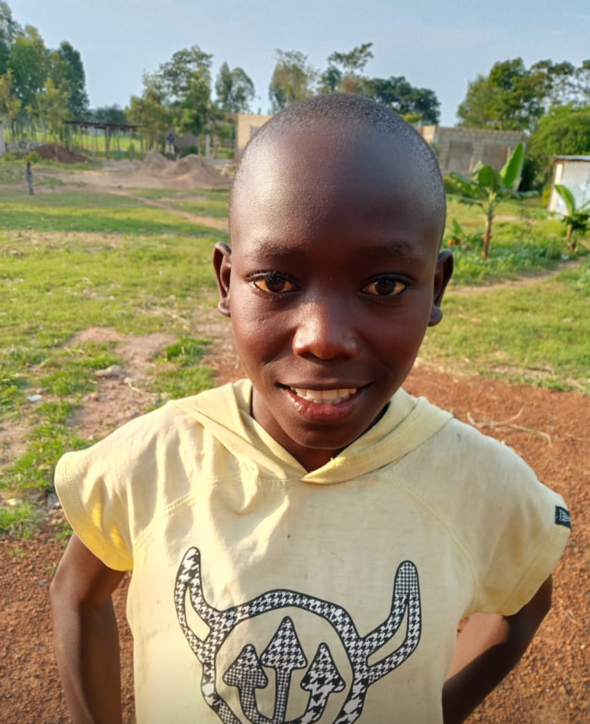 Young African boy smiles outdoors, wearing yellow hoodie, with green grass and buildings in the background.