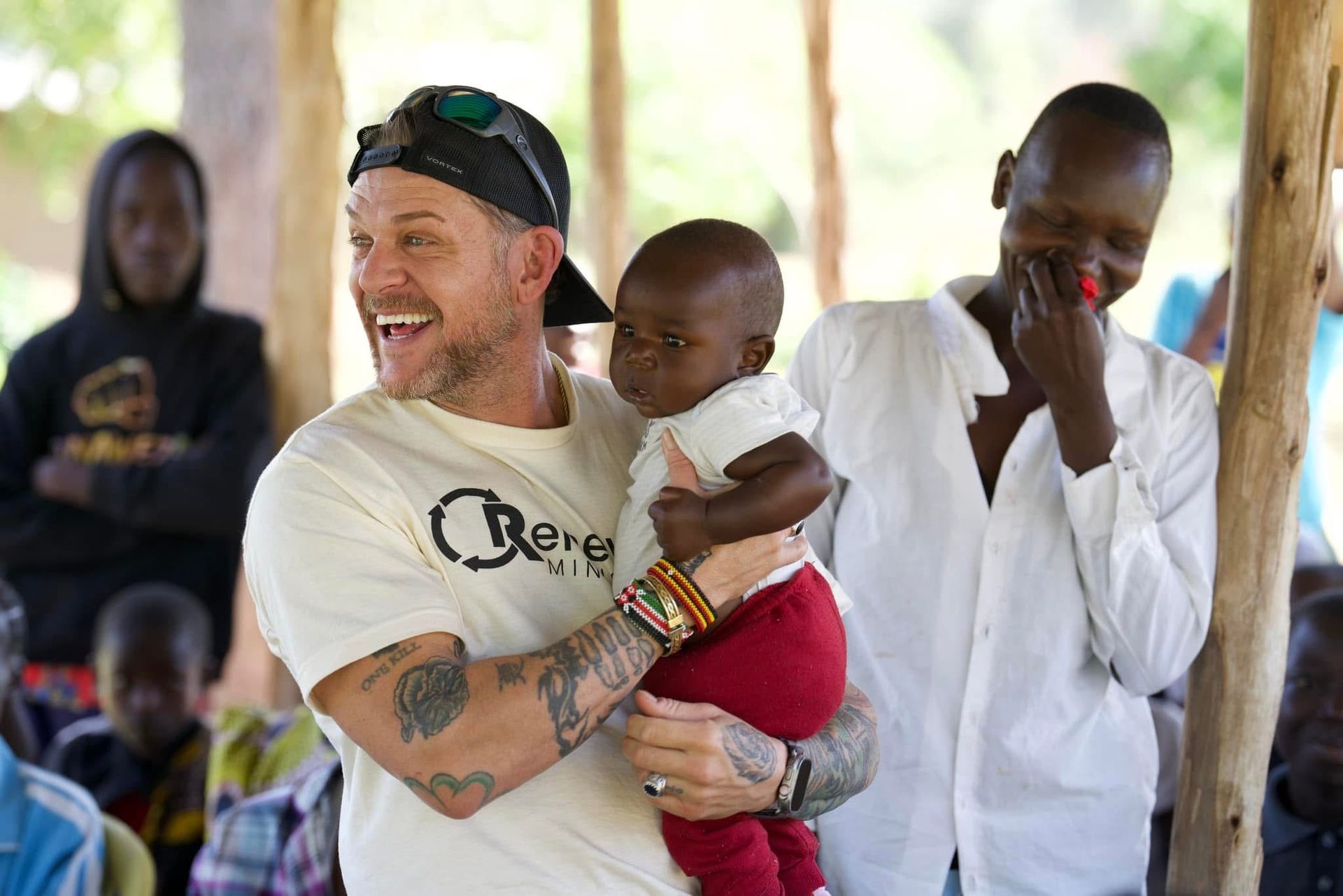 Man holding baby smiles, surrounded by people in a sunny outdoor setting.
