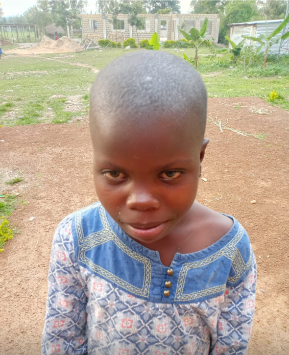 Young child with shaved head, wearing patterned blue and floral top, looking directly at the camera, outdoors.