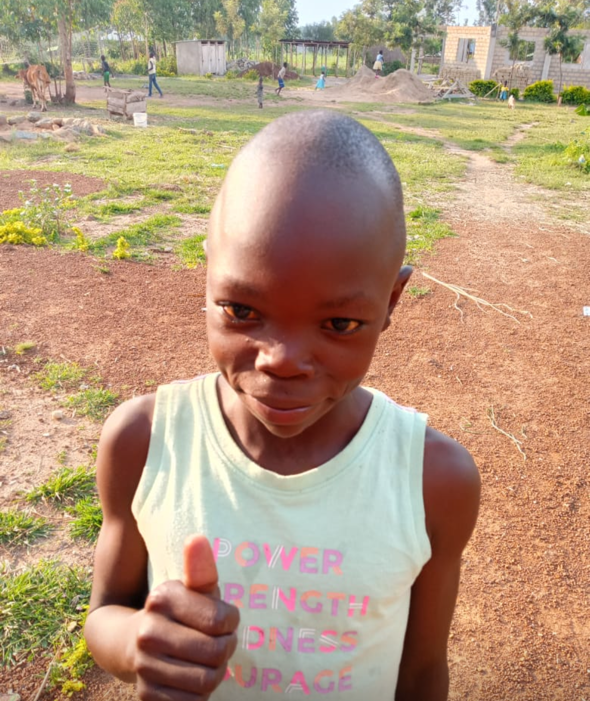 Young person with shaved head and thumbs up, smiling; outdoors.
