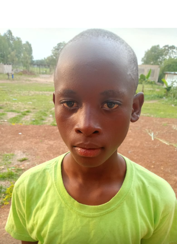 Young Black boy with shaved head in a green shirt stands outdoors, looking at the camera.