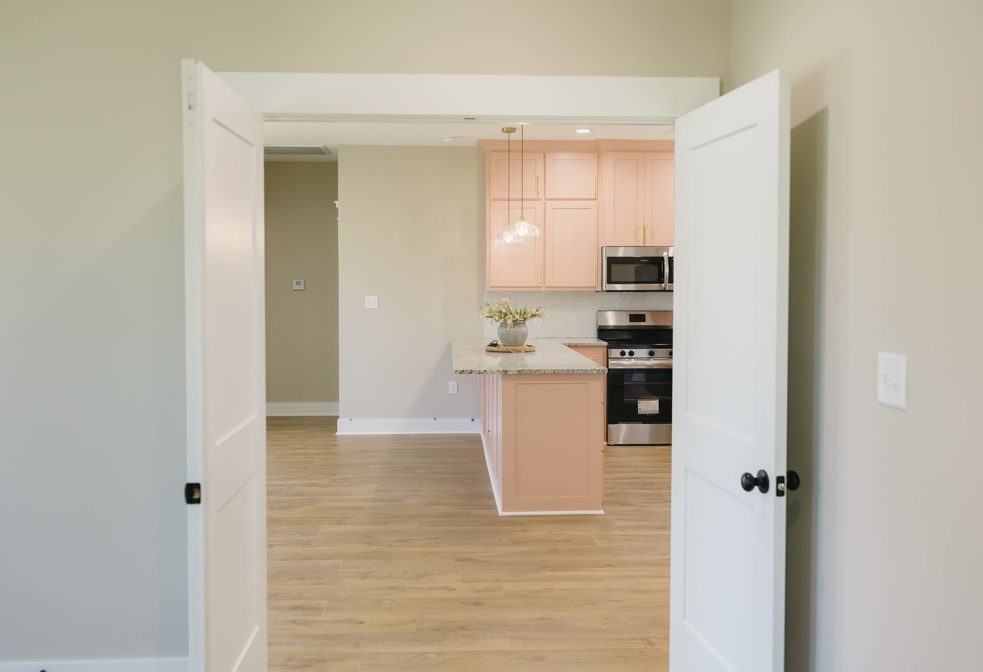 Open white double doors framing a kitchen with light pink cabinets and wood floors
