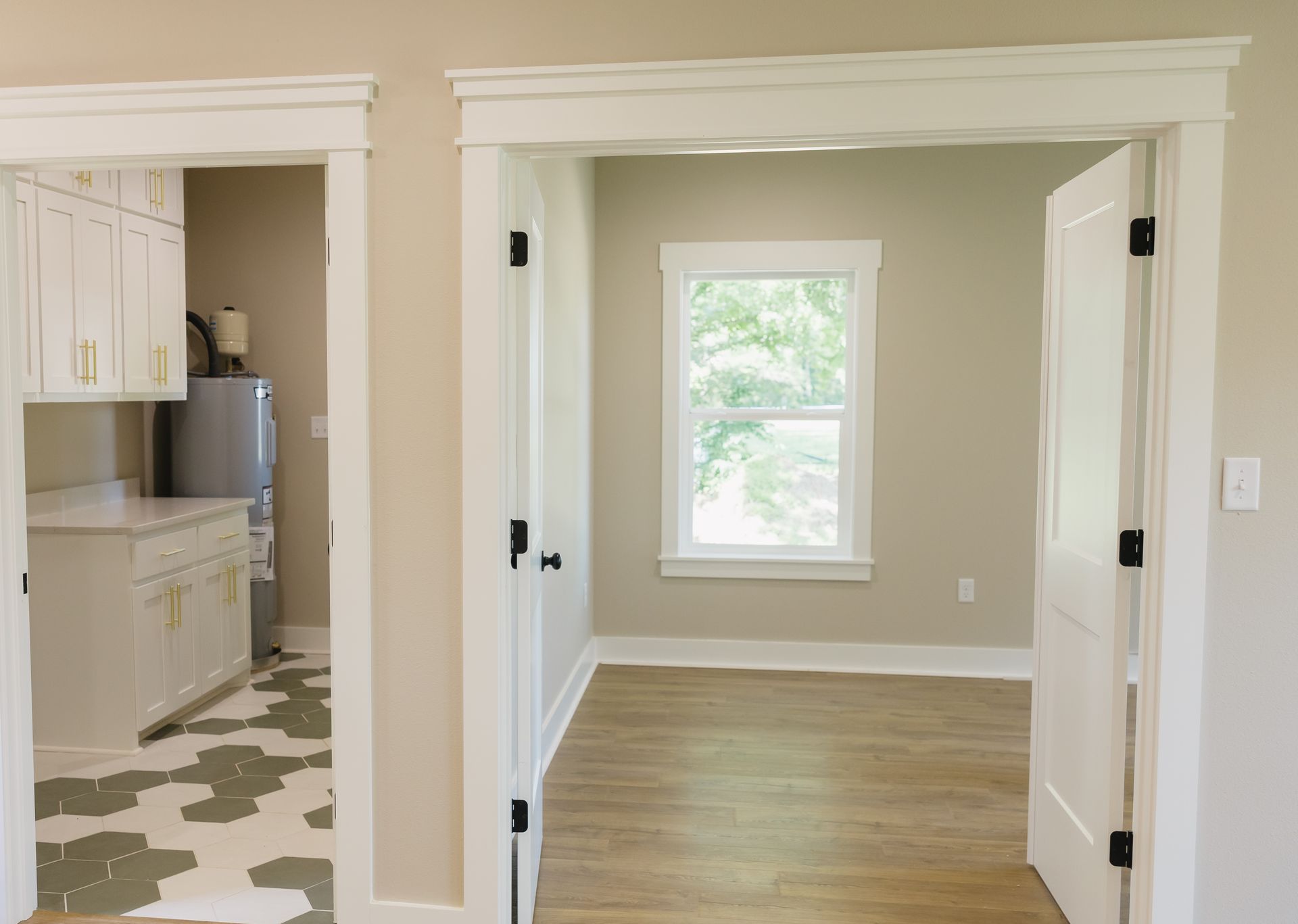 Empty room with wood floor and window, viewed through open doorway beside a kitchen with patterned tile floor