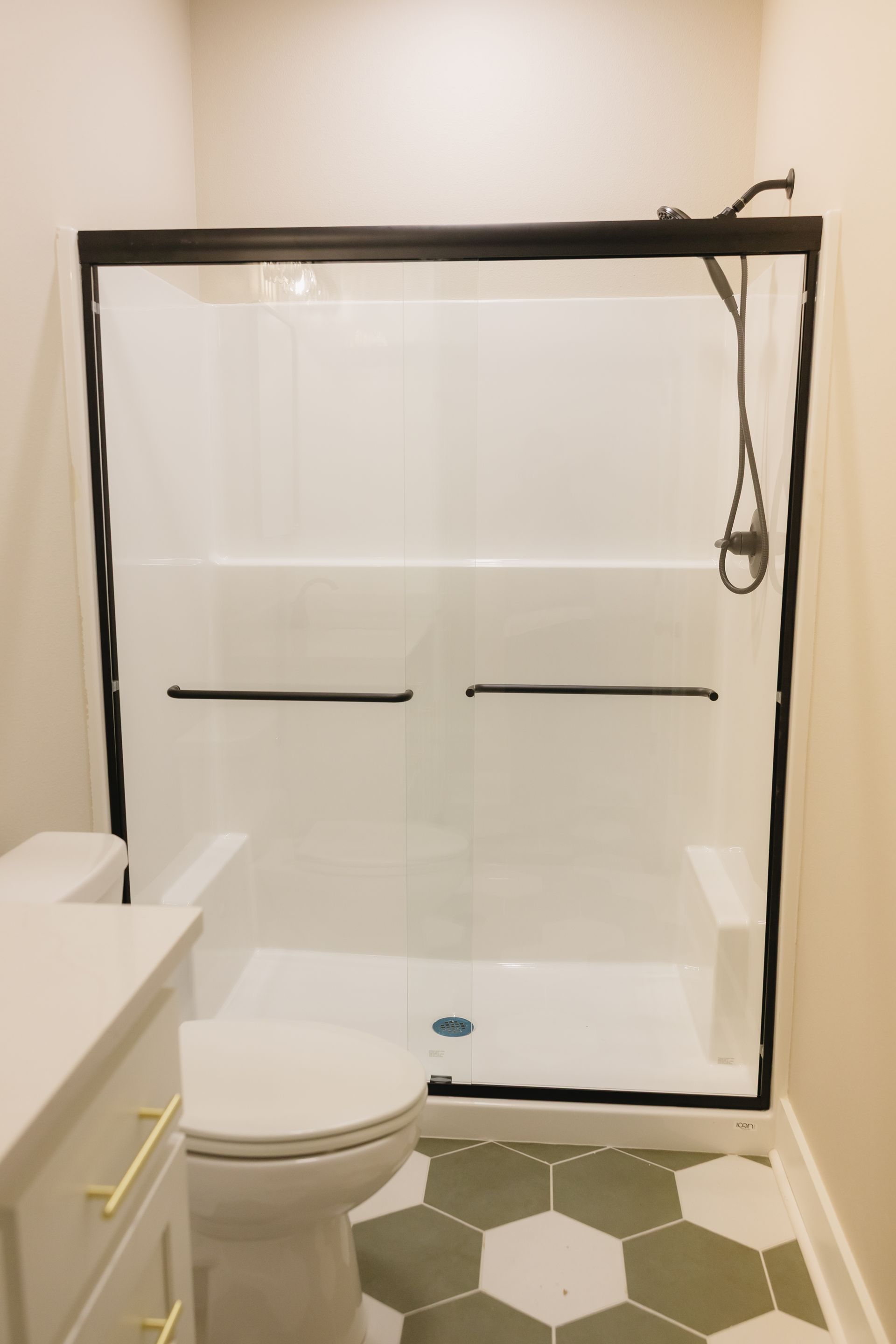 Small bathroom with black-framed shower door, white tub, toilet, and green hexagon floor tiles.