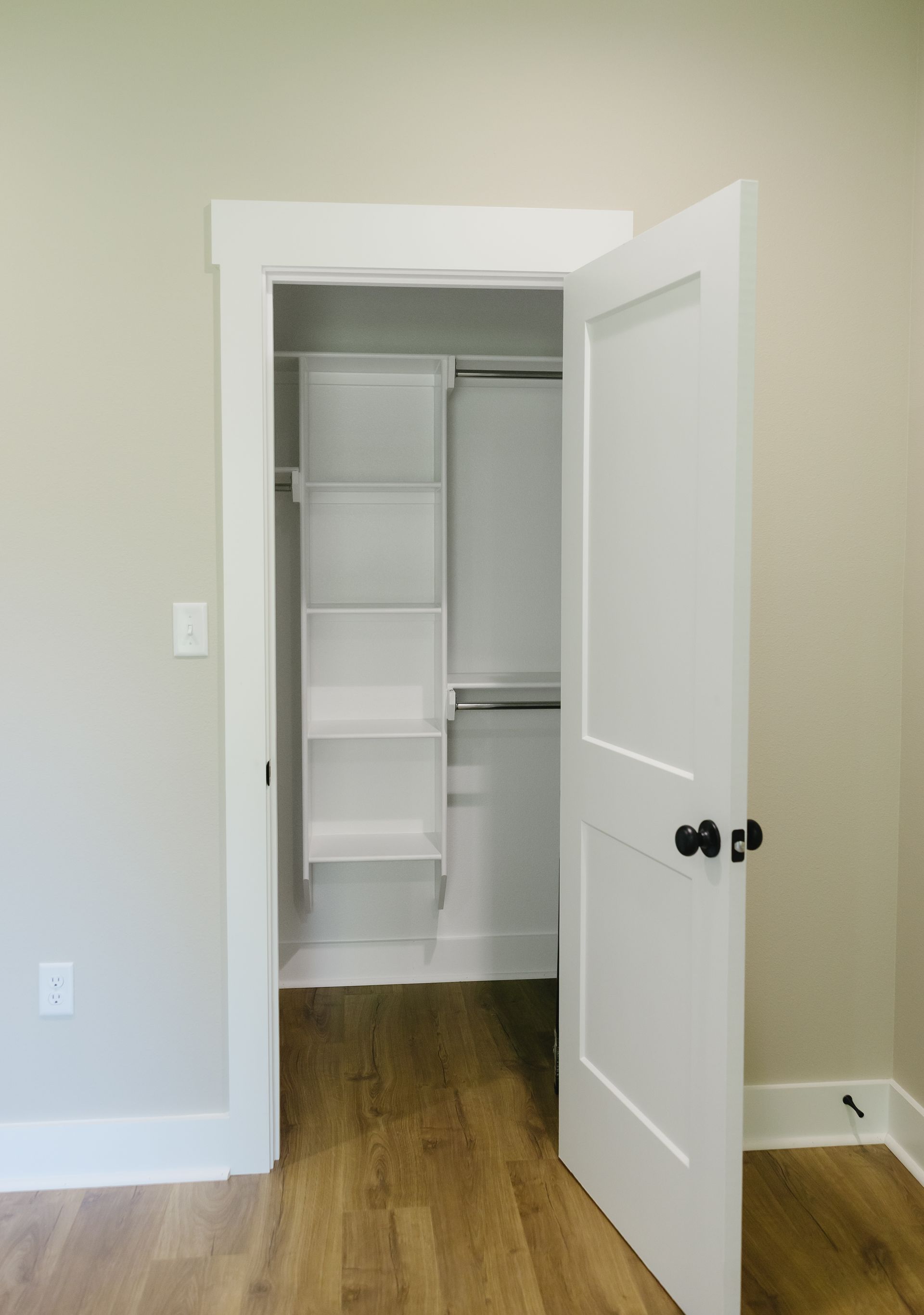 Open white closet with shelves and hanging rod in a beige room with hardwood floor.