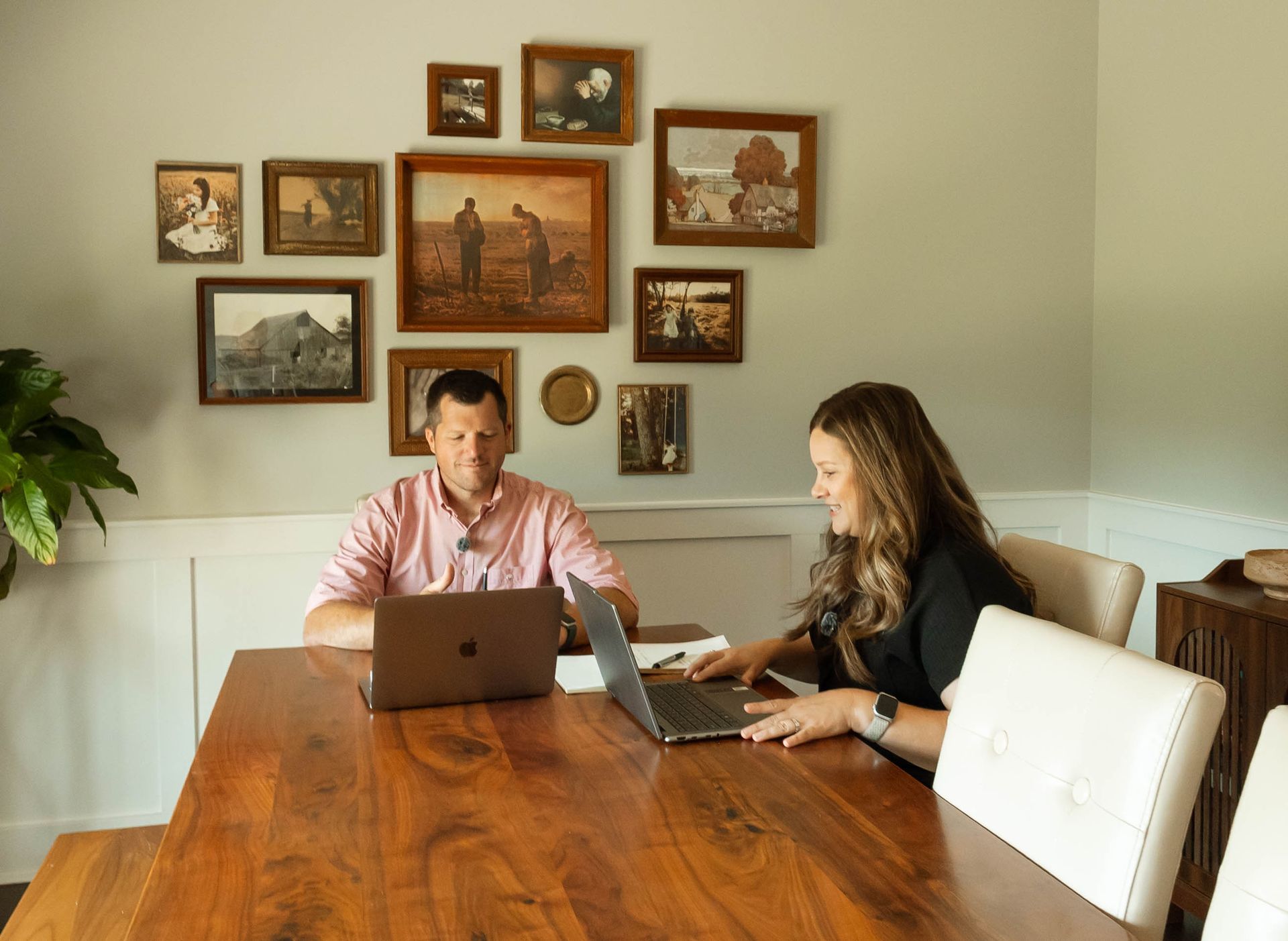 Two people working on laptops at a wooden dining table in a bright room with framed wall art.