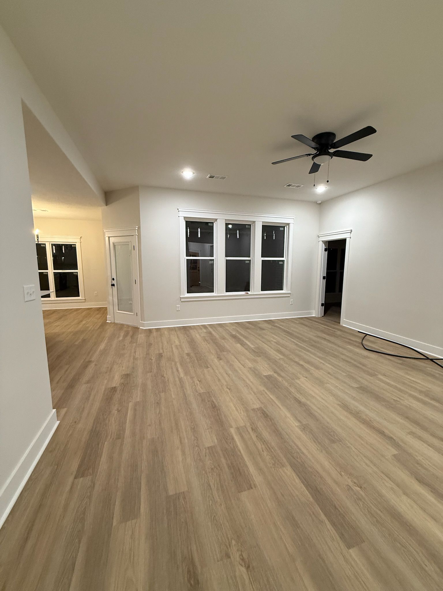 Empty modern living room with light wood floors, white walls, ceiling fan, and large windows.