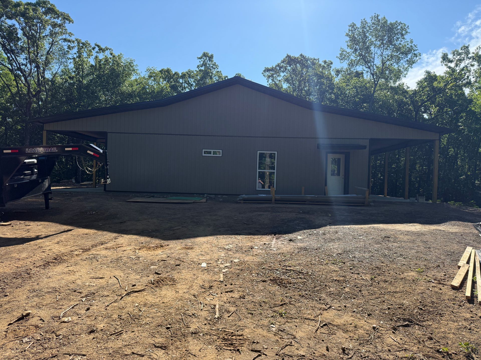 Gray metal building under construction with bare dirt foreground and trees around it