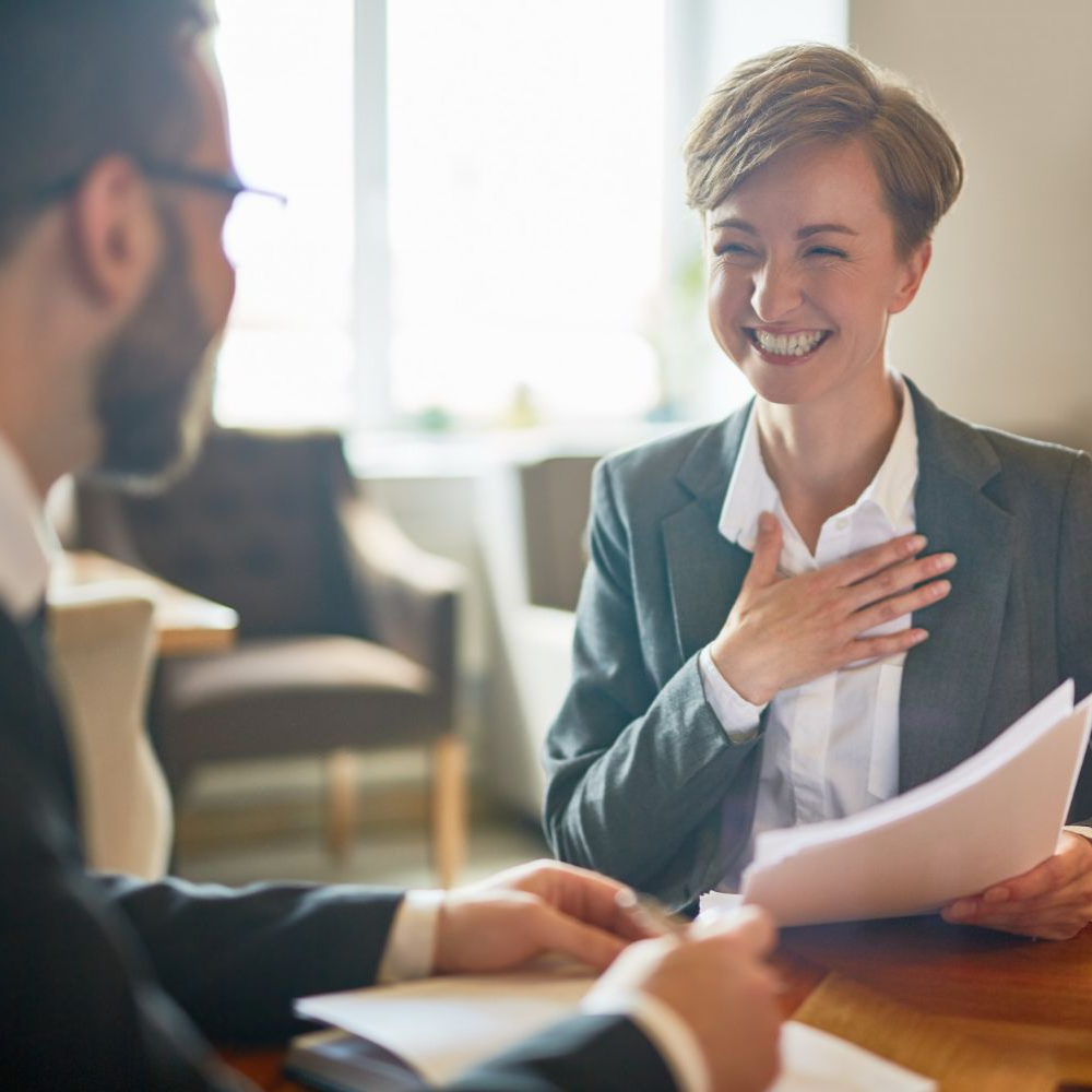 Two coworkers smiling during a business meeting, one holding papers at a table in a bright office.