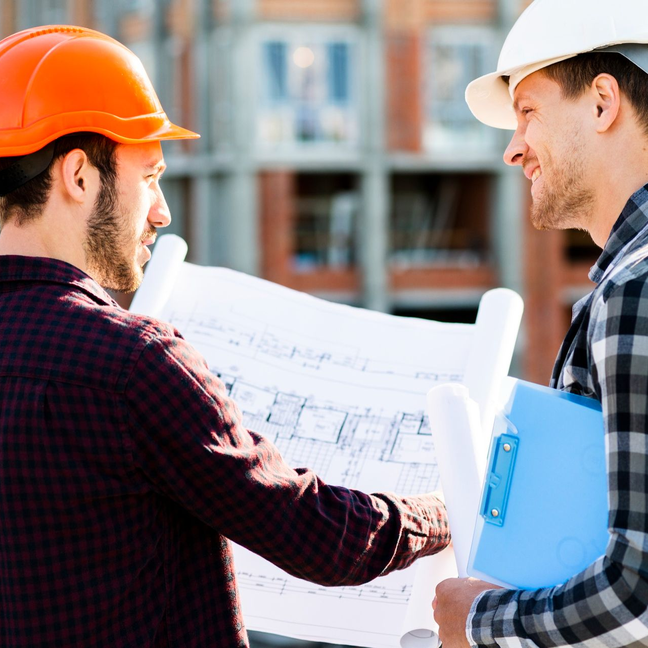 Two construction workers reviewing blueprints at a building site, one in an orange hard hat.