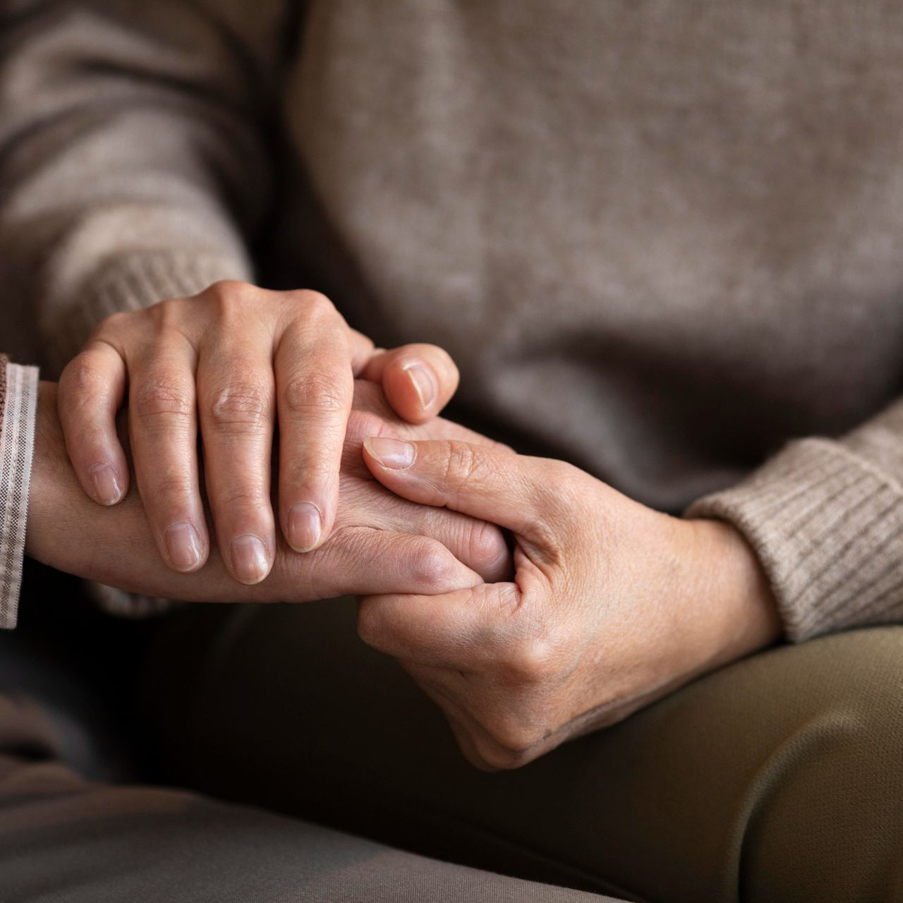 Two hands gently clasped on a lap, wearing beige sweaters in a soft indoor setting