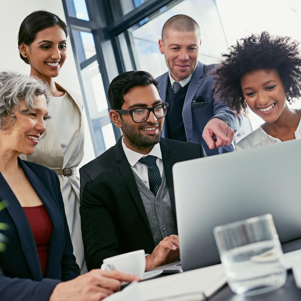 Colleagues gathered around a laptop in a bright office, smiling and discussing work.