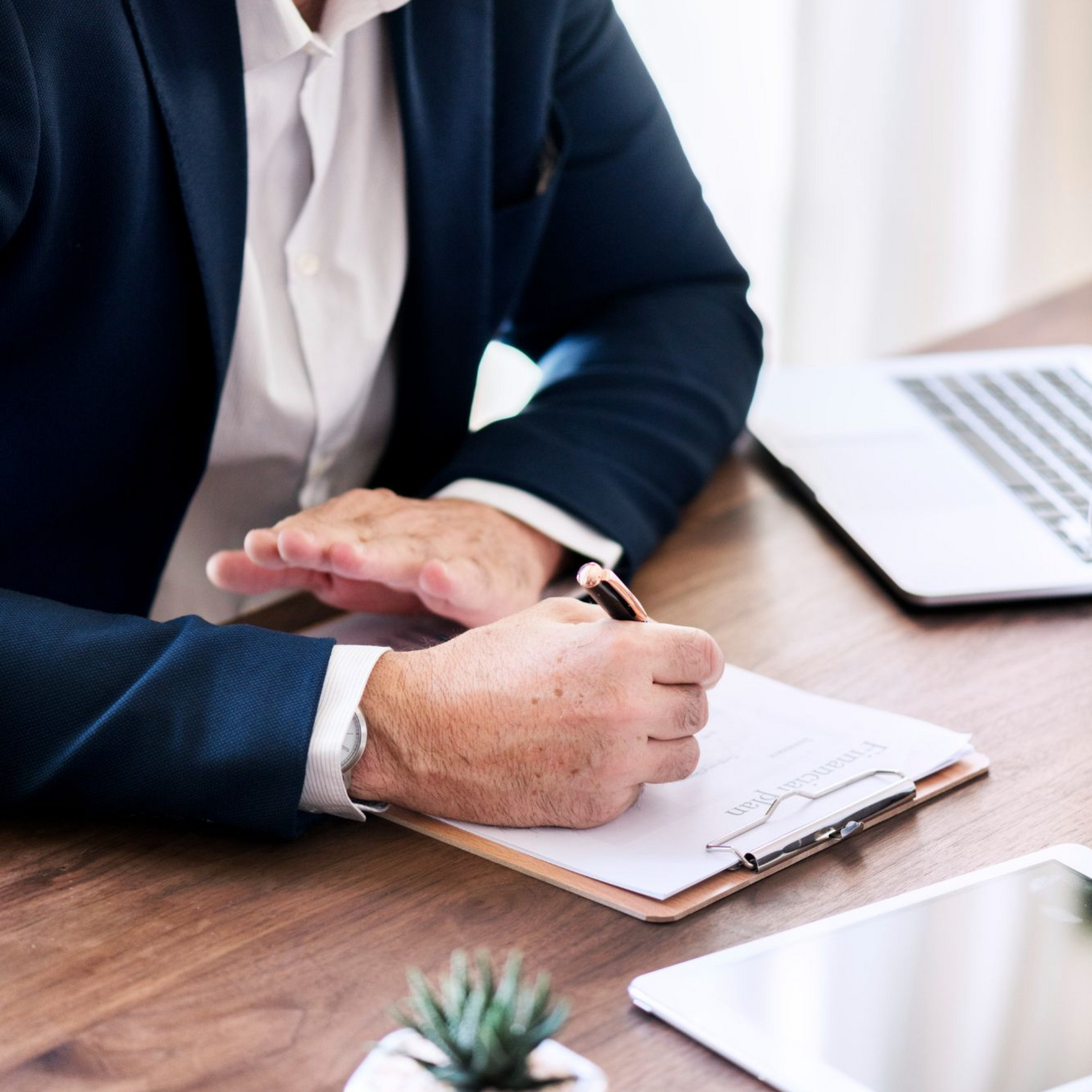 Person in a navy blazer writing on a clipboard at a wooden desk beside a laptop and tablet
